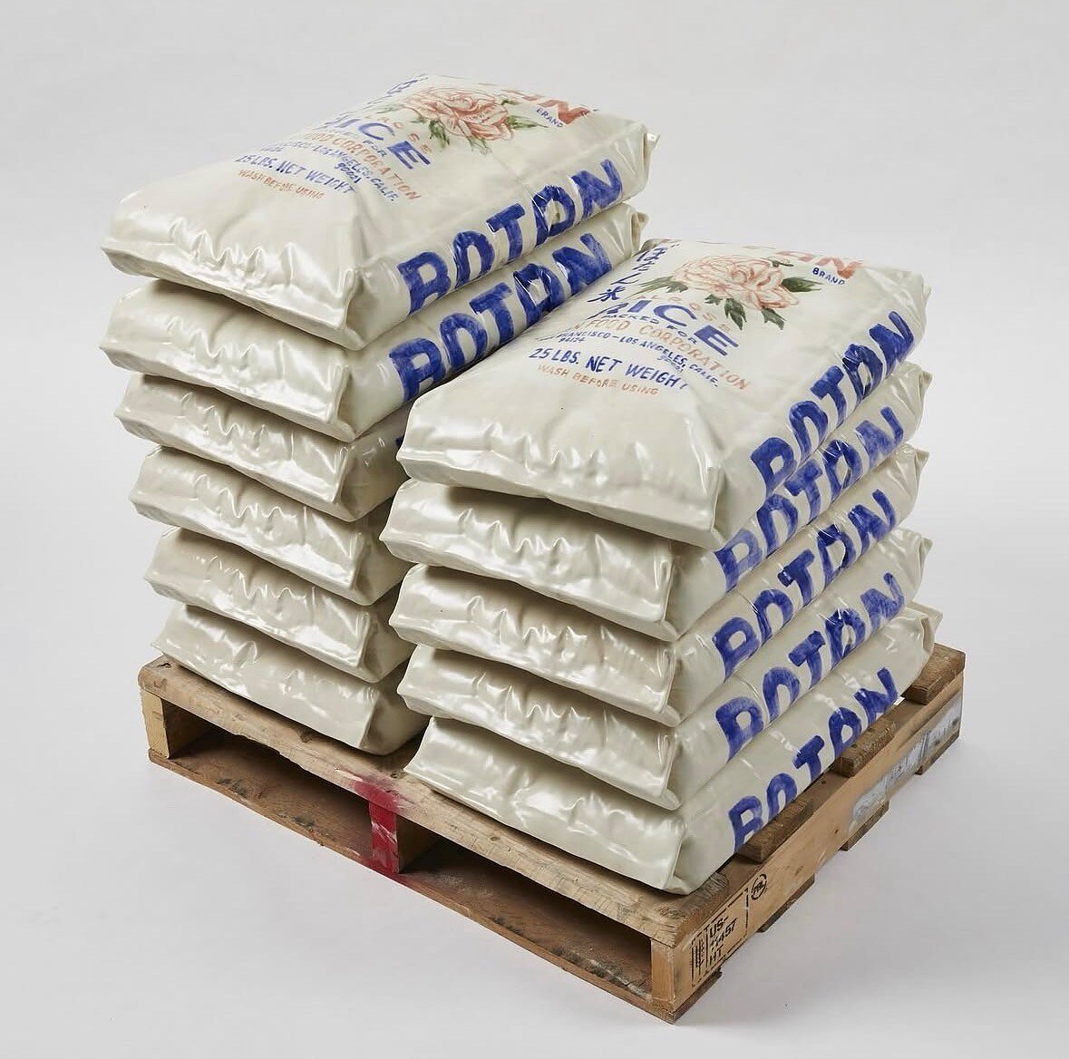 A highly detailed ceramic sculpture of ten Botan Rose-brand rice bags on a wood pallet.