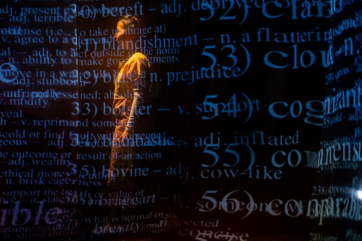 A woman is on stage, illuminated by yellow light with a flurry of words and definitions in blue projected on top of her on transparent screens. 