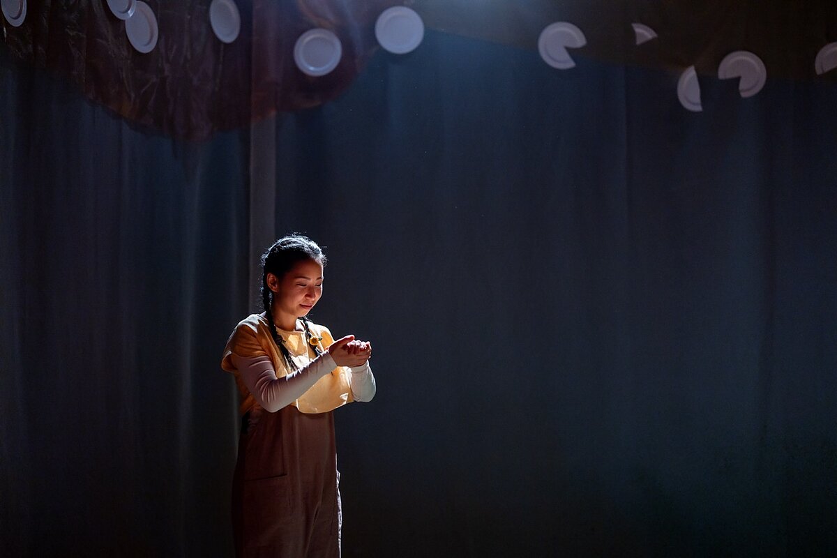 A smiling woman on stage stands with her hands clasped together. Above her are white dinner plates, some chipped. 