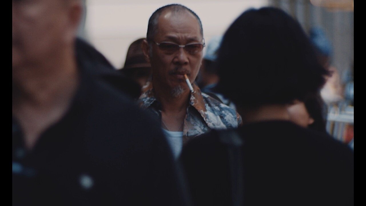 A man wearing sunglasses and smoking a cigarette moves through a busy city street.