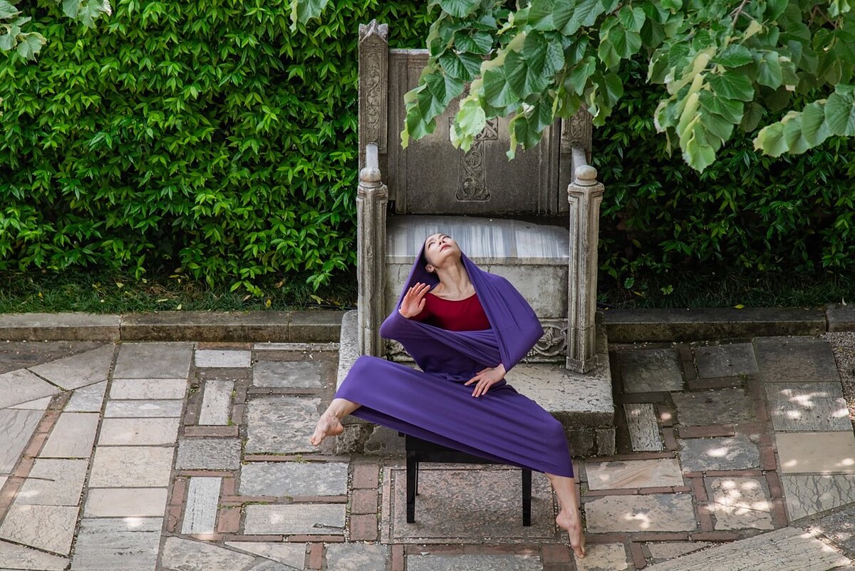 A woman dancer seated outdoors in a courtyard dances, appearing to emerge out of a purple shroud. 