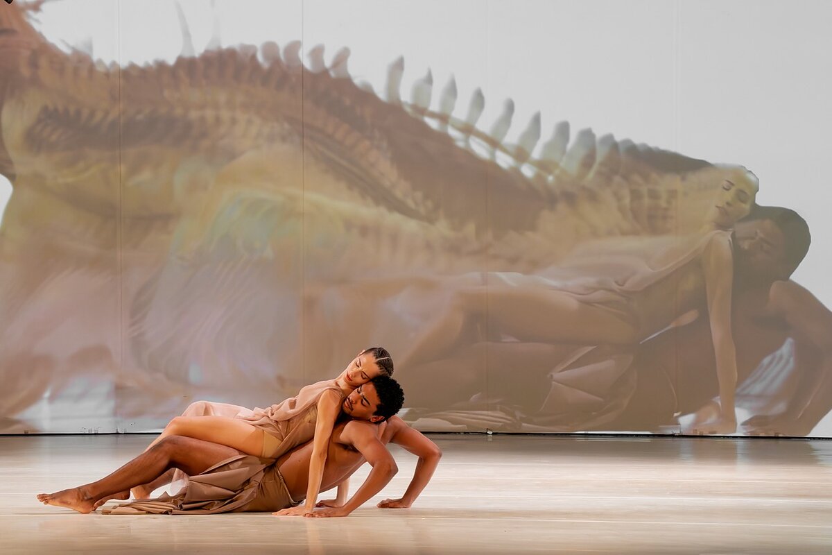 Two dancers on stage recline on top of one another while a projection in the background features their images duplicated.  