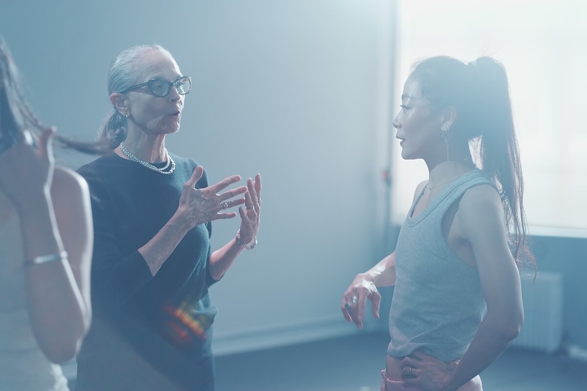 a woman on the left with white hear wearing glasses speaks to a woman on the right in a sunlit room. 