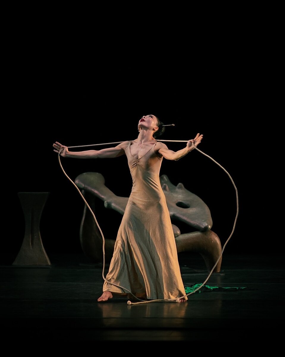 A woman dancer on stage wearing a beige dress and holding a cord across her shoulders as she looks up. 