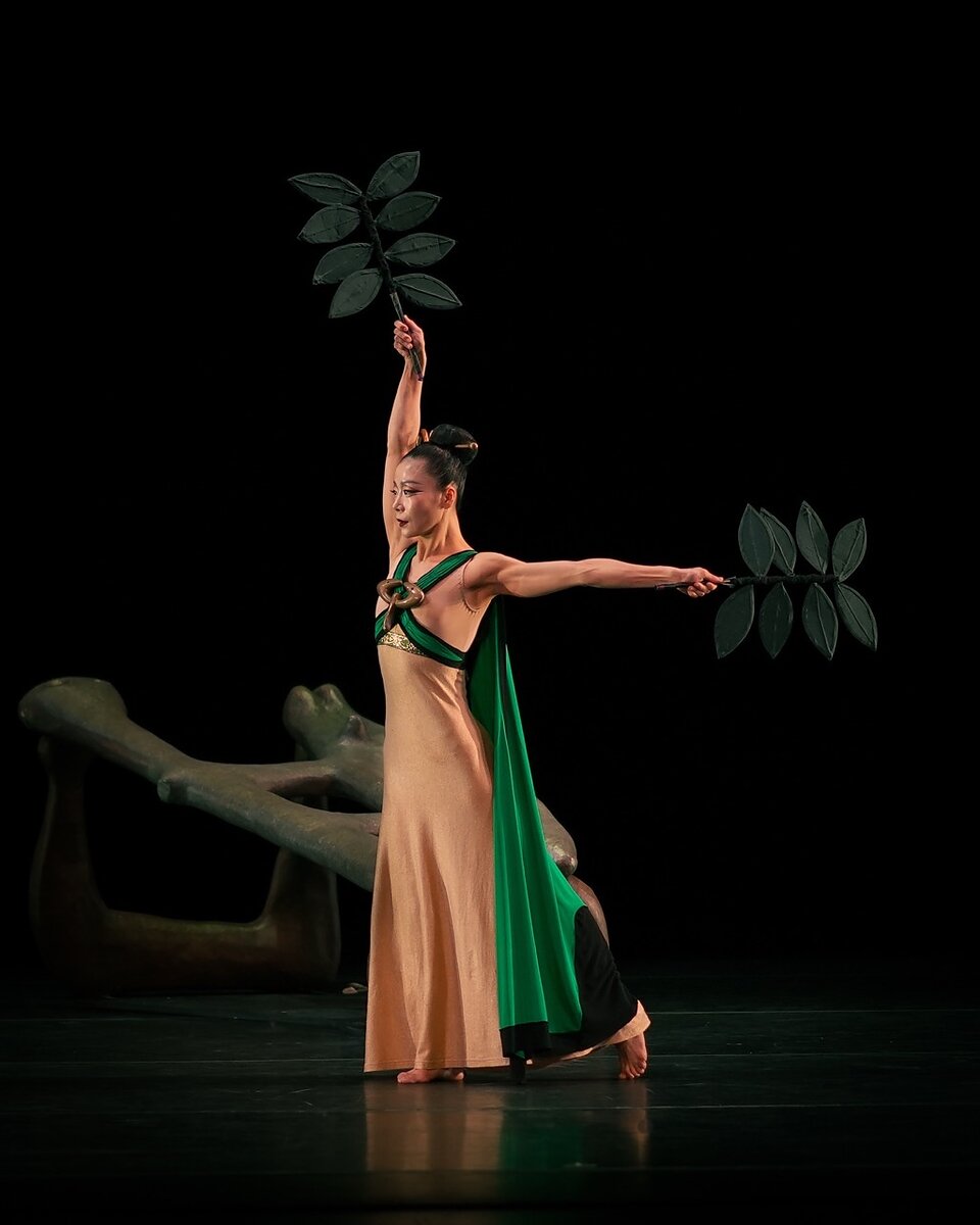 A woman dancer on stage wearing a peach colored dress with a green cape holds up leaf-shaped props. 