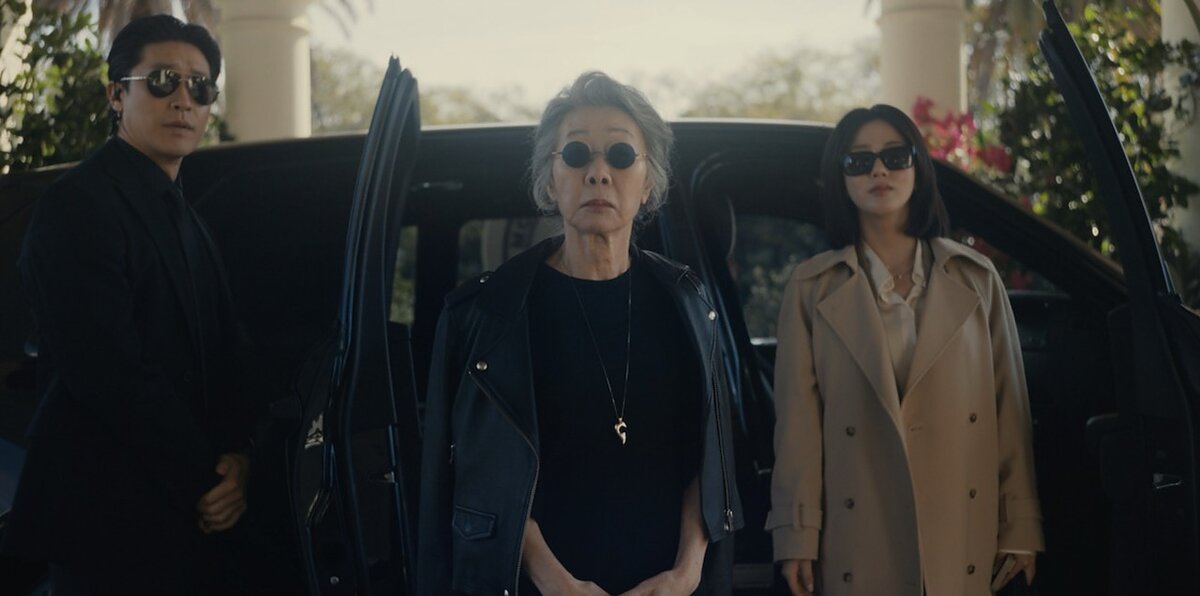 An elegant older woman dressed in black and wearing sunglasses exits a vehicle with two younger associates flanking her. 