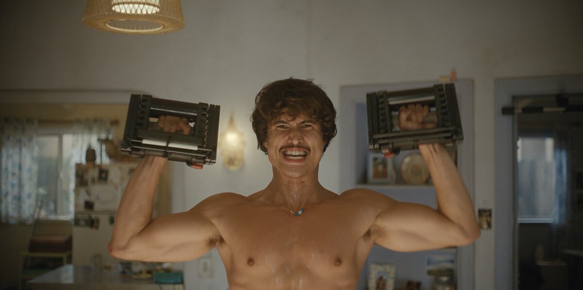 A shirtless young man grimaces while lifting weights in a living room. 
