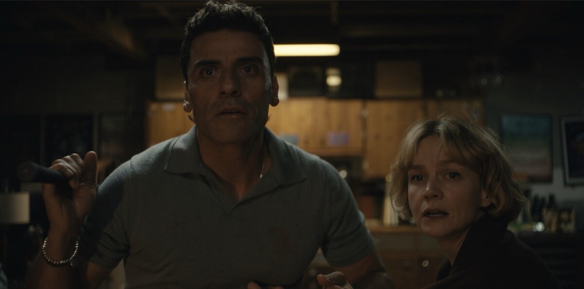 A man and a woman in a dimly lit room look anxiously at the camera. 