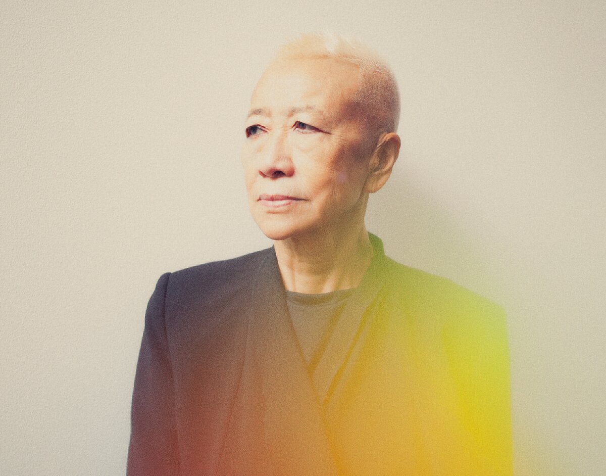 An older Asian woman with a blonde buzz cut wearing a black blazer is seen in three-quarter profile, looking to the left. There is a glow of orange and yellow light from the camera.  