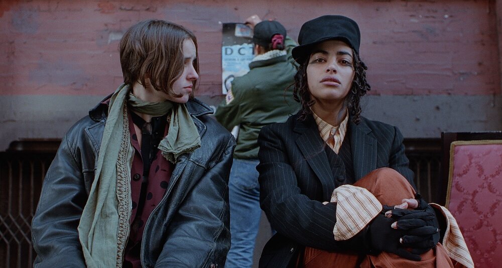 A woman with short hair looks at the woman sitting next to her who stares at us. Behind them is someone hanging up a poster on a brick wall. 