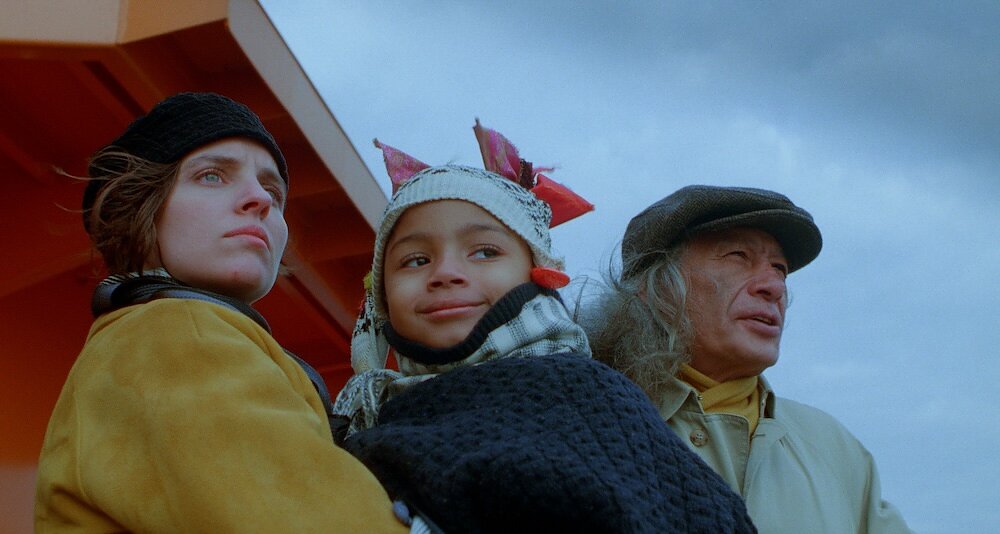 A child wearing a knit hat smirks while looking off in the distance. She is held by a woman in a yellow jacked and beret who also looks out with a serious expression. They are standing next to an older man in a news cap. 