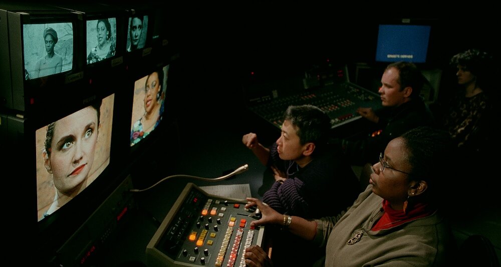 Three people sit in front of a control room desk watching a series of television monitors showing a talk show. 
