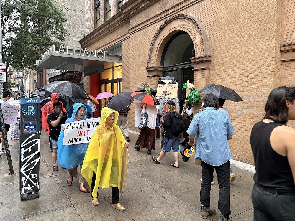 Protesters wearing rain ponchos and holding umbrellas and signs parade in front of a brick building.