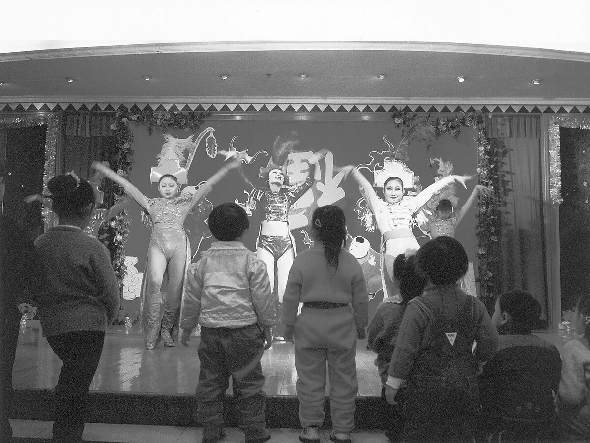 A black and white still of young children gazing at performers on a low stage. 