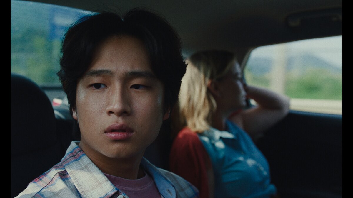 A close up of a worried young man who looks out of a car window while sitting next to a young woman. 