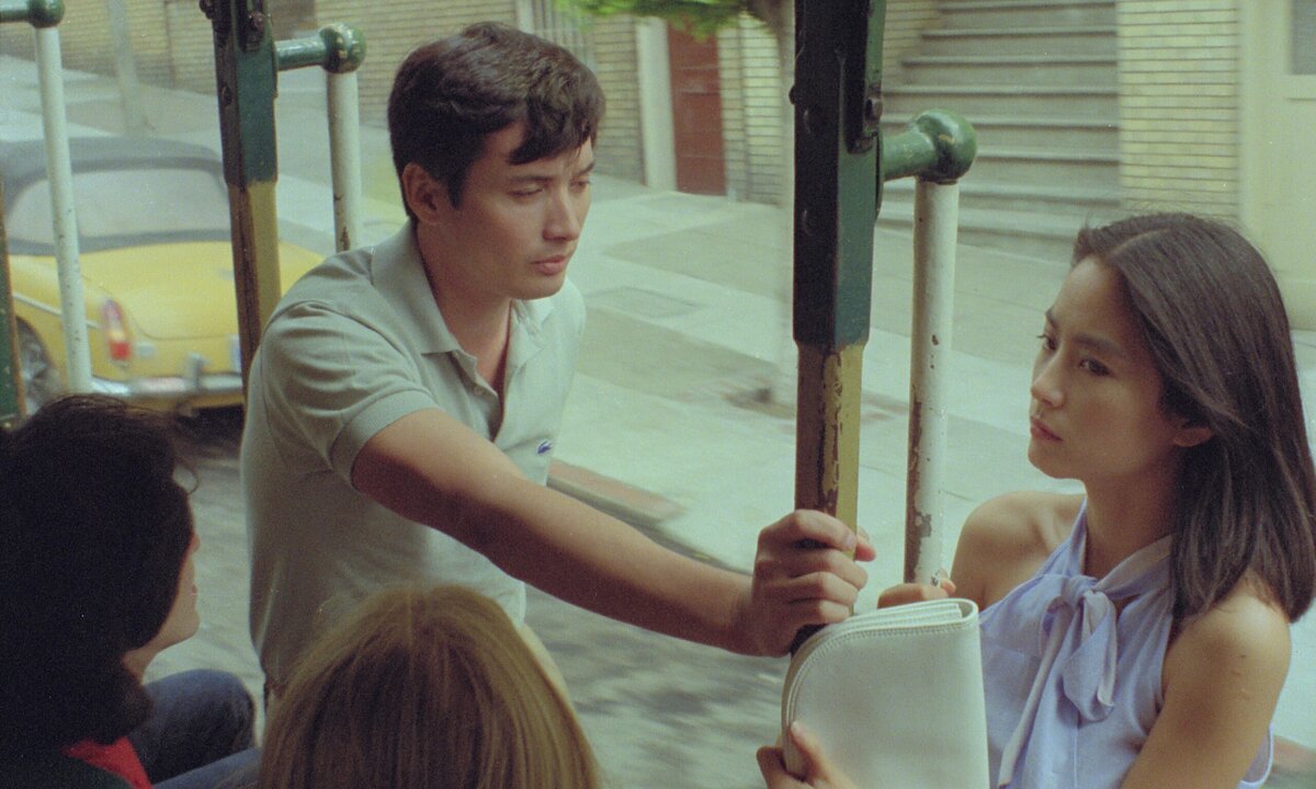 A man gazes seriously at a woman looking away while leaning on a trolley pole. 