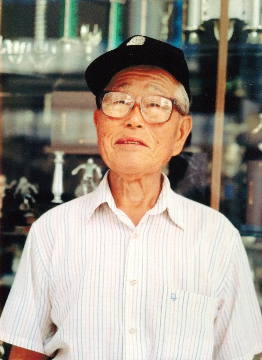 An elderly man wearing a baseball cap and glasses stands in front of a trophy case.