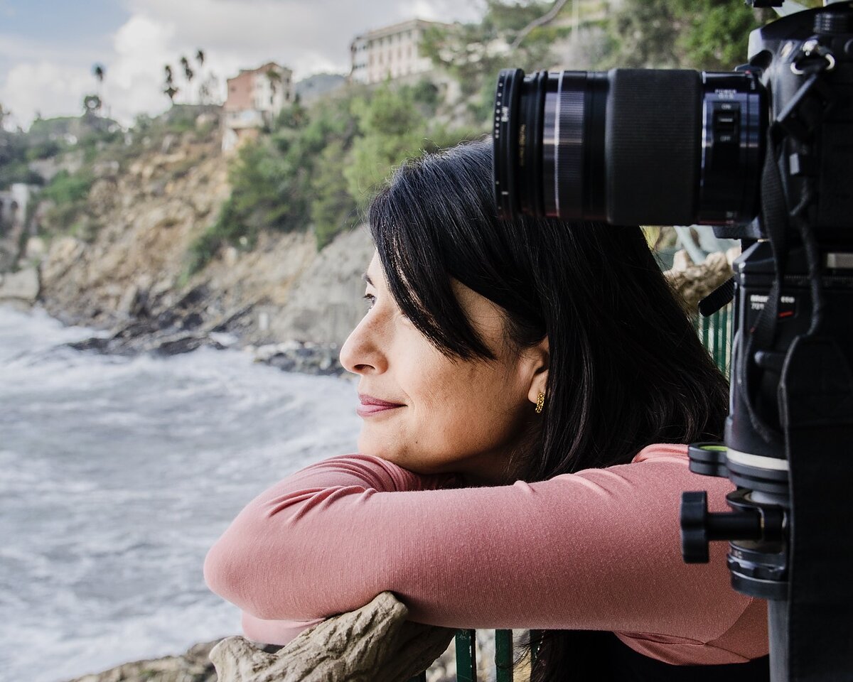 A woman next to a camera leans over a railing looking out onto the ocean.