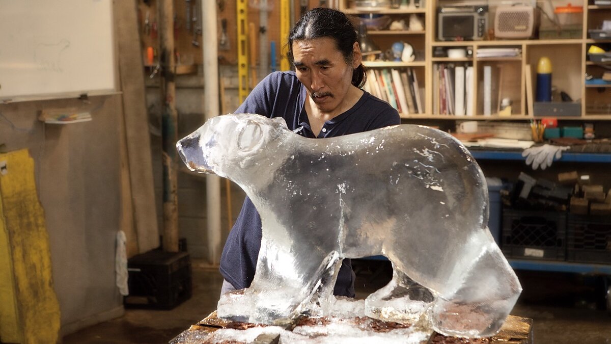 A man in a workshop carves an ice sculpture of a polar bear cub.