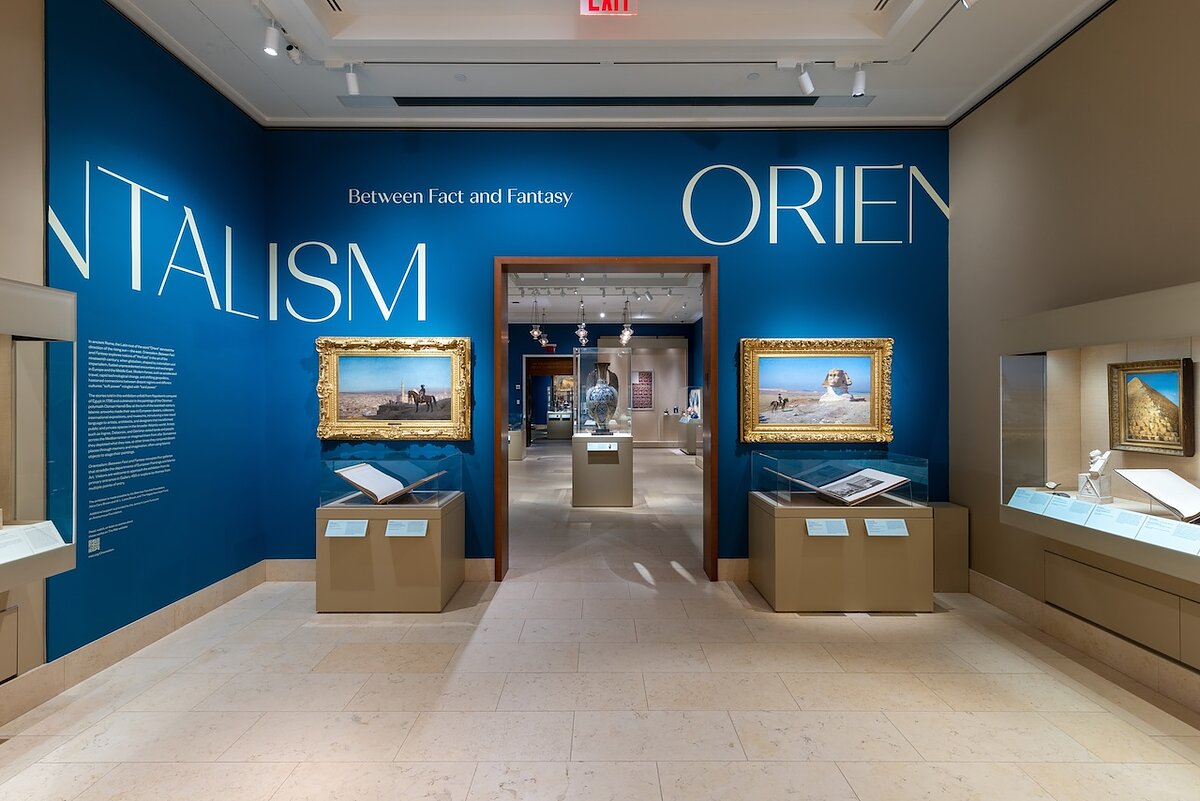 An art gallery with the words "Orientalism: Between Fact and Fantasy" written on blue walls and glass-covered display cases and paintings with ornate gold frames. 