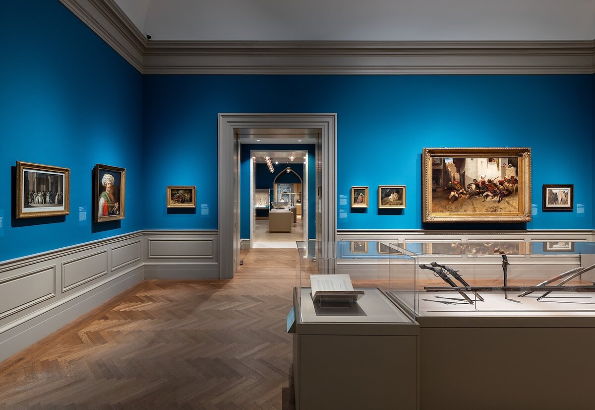A gallery with blue walls filled with paintings and glass vitrines.