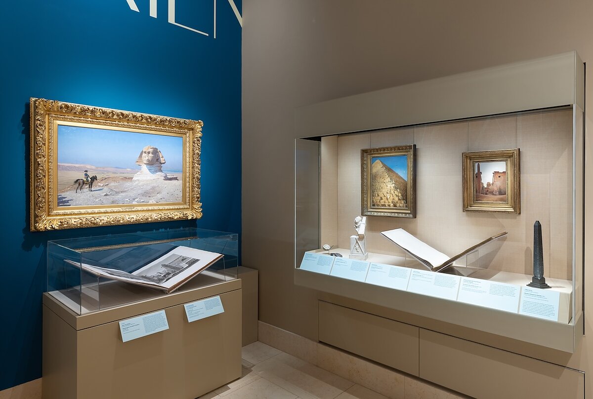 A gallery corner with blue and tan walls displays old paintings and books in glass display cases.