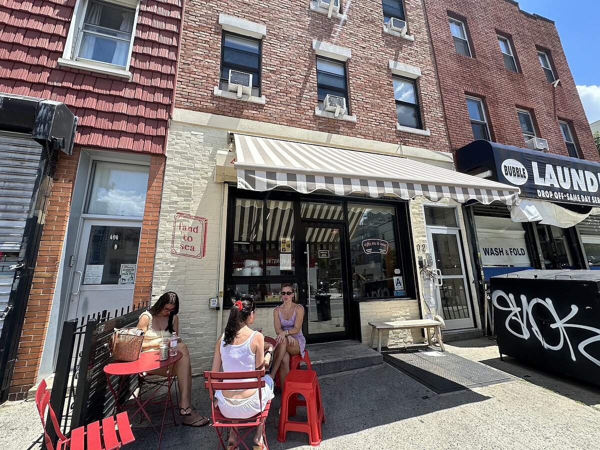 People seated on red chairs outside a small cafe with a beige and white striped awning on a sunny summer day. 