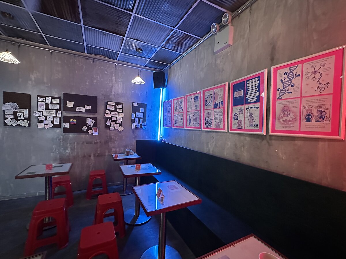 Colorful artwork hangs on the walls of a cafe lit by a neon blue line with red plastic chairs and grey walls. 