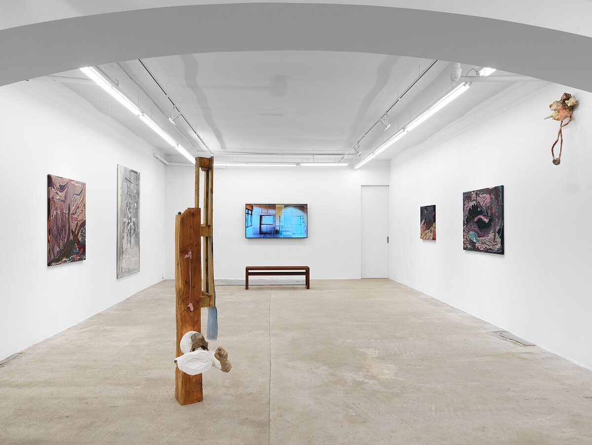 Installation image of a gallery featuring two paintings, a video work, and several sculptures on the floor and hung on the wall. 