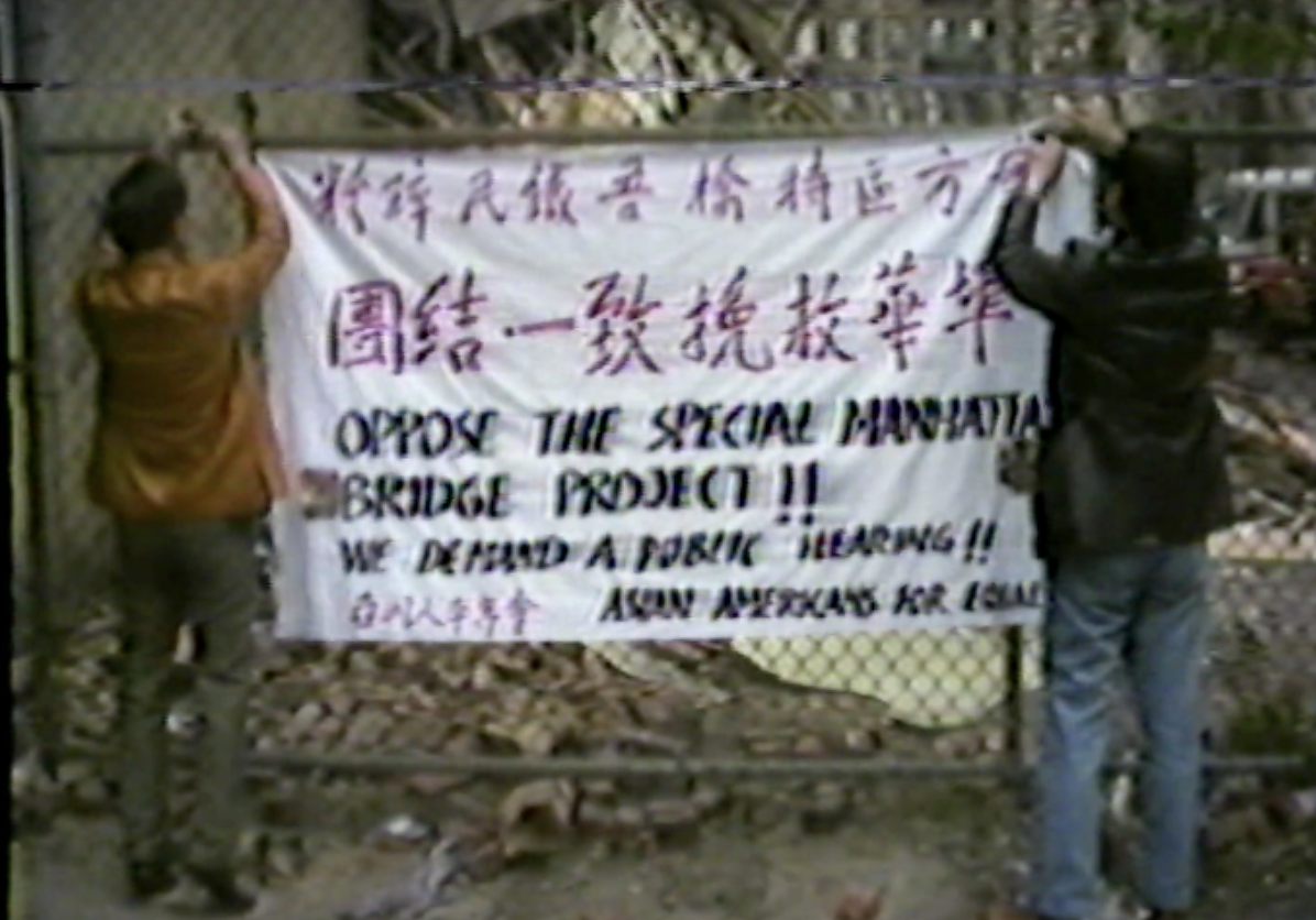 Grainy footage showing two men hanging a banner that says "Oppose the special Manhattan Bridge Project!!" in English and Chinese onto a chainlink fence. 