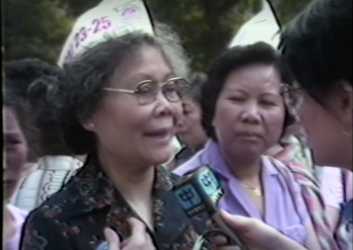Grainy footage of an older woman wearing a paper hat being interviewed by a television reporter in front of other women. 