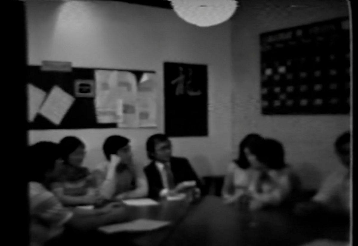 Grainy black and white footage of a group of people sitting at a table. 