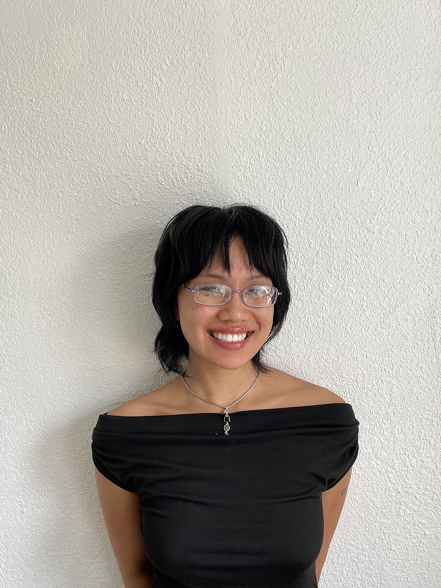 an Asian person wearing metal rimmed glasses and an off-the-shoulder black top smiles against a textured white background