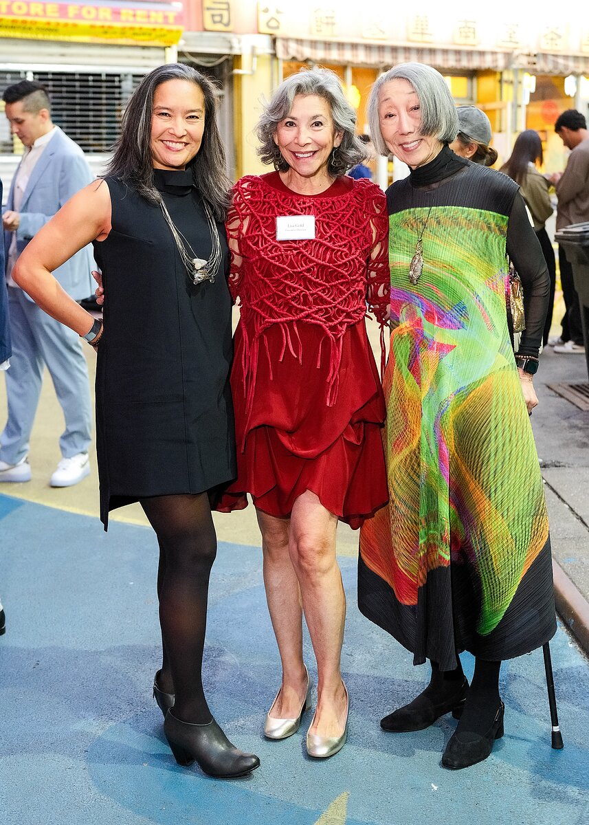 Three women pose together on a bustling and colorful street.