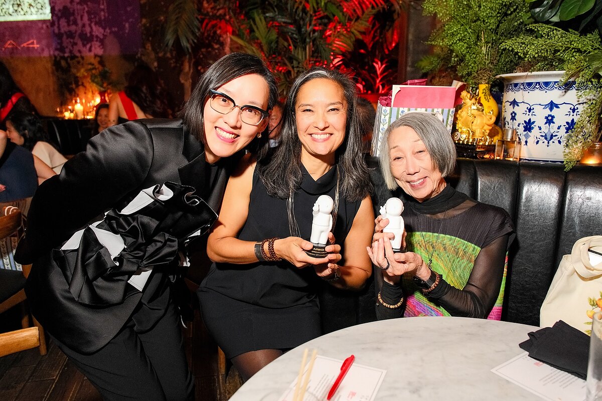 Three women post together while seated at a booth. Two are holding awards.