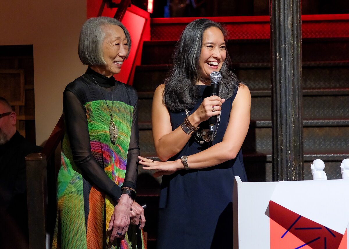 Two women stand together at a podium smiling. One is speaking into a microphone.