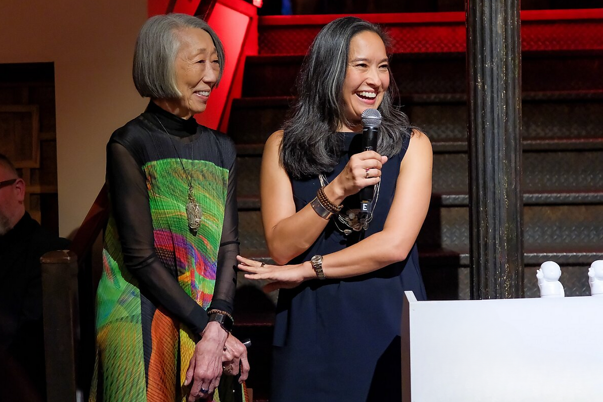 Two women stand together at a podium smiling. One is speaking into a microphone.