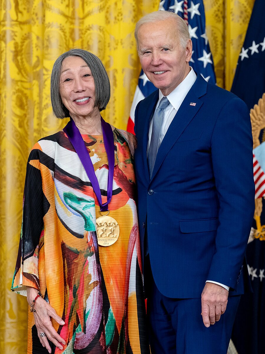 A woman wearing a large golden medal stands next to a man wearing a blue suit in front of a gold backdrop and American flag.