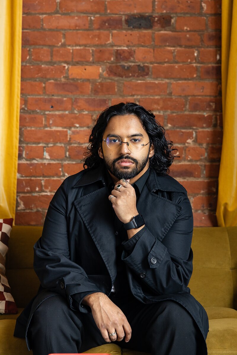 a man wearing a black coat with large lapels sits on a couch and holds one hand to his chin