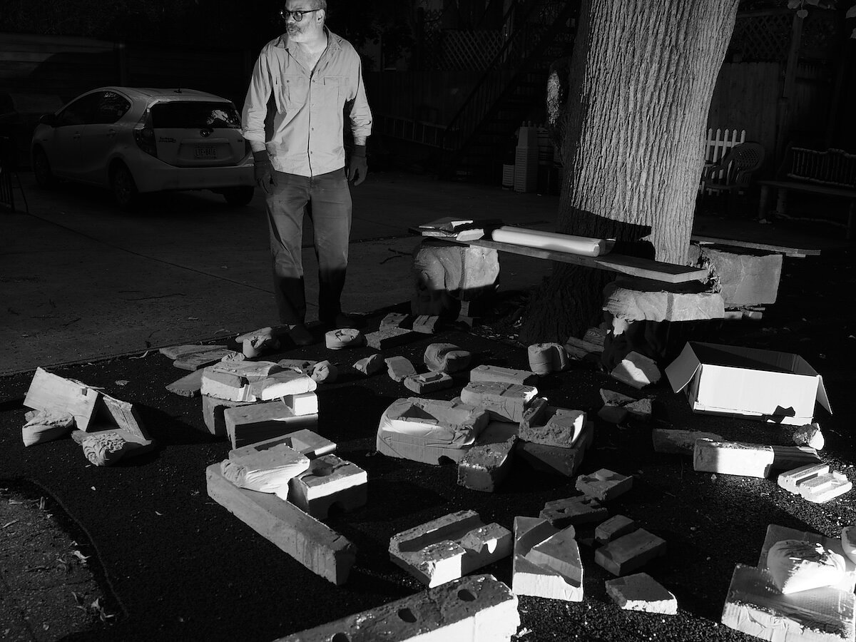 A black and white photograph of a man standing outside in glasses and black gloves in front of a pile of concrete shapes. 