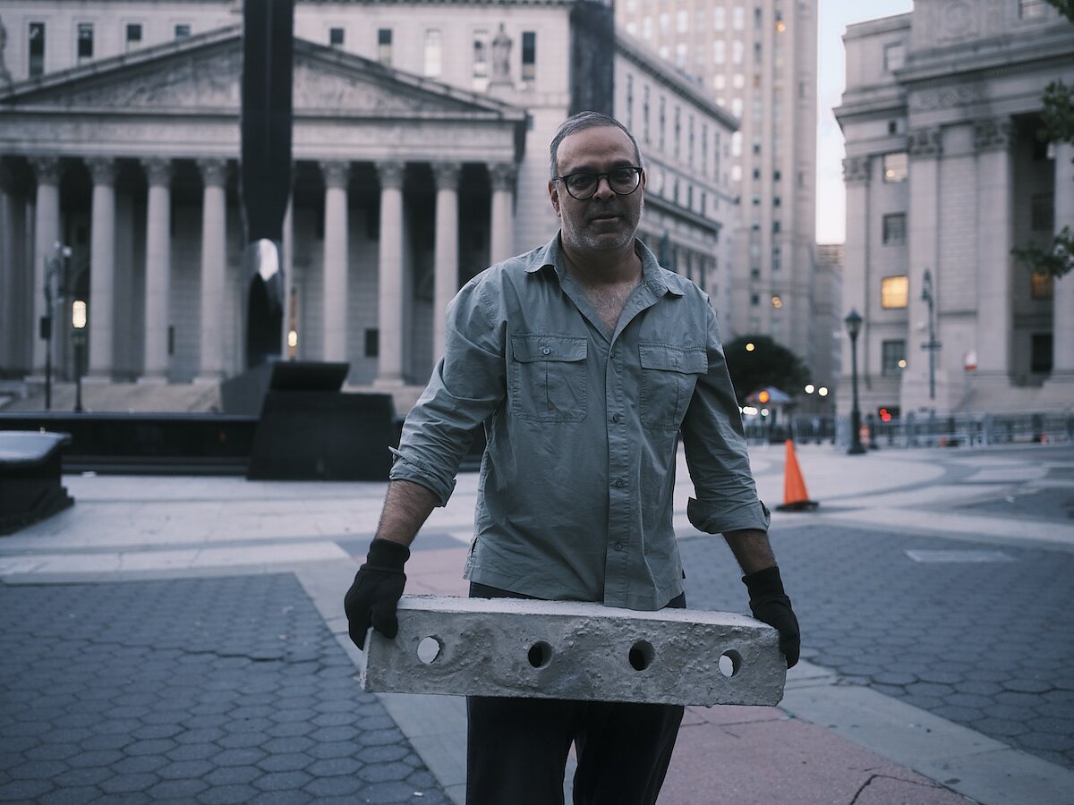 A man in glasses and wearing black gloves stands in the middle of a town square holding what appears to be a long cement block. 