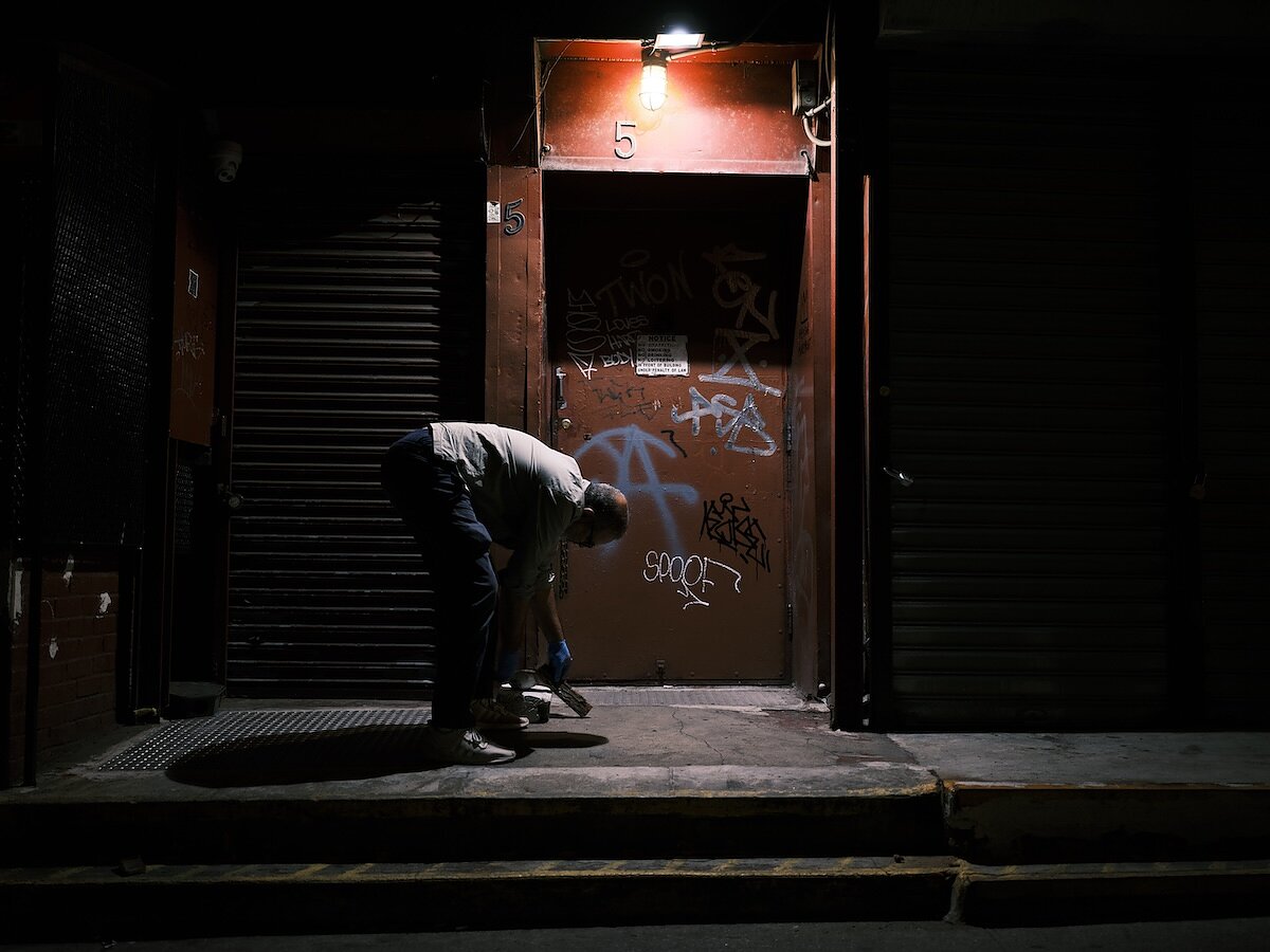 A man hunches over at night to put something on the ground in front of a city building with graffito on it. 
