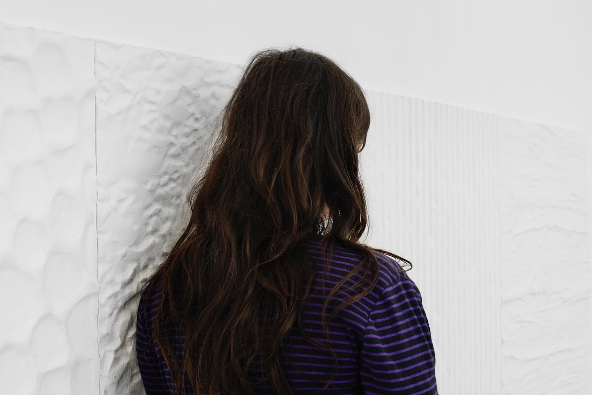 A woman with long brown hair is seen from behind rubbing against a white wall of textured surfaces.