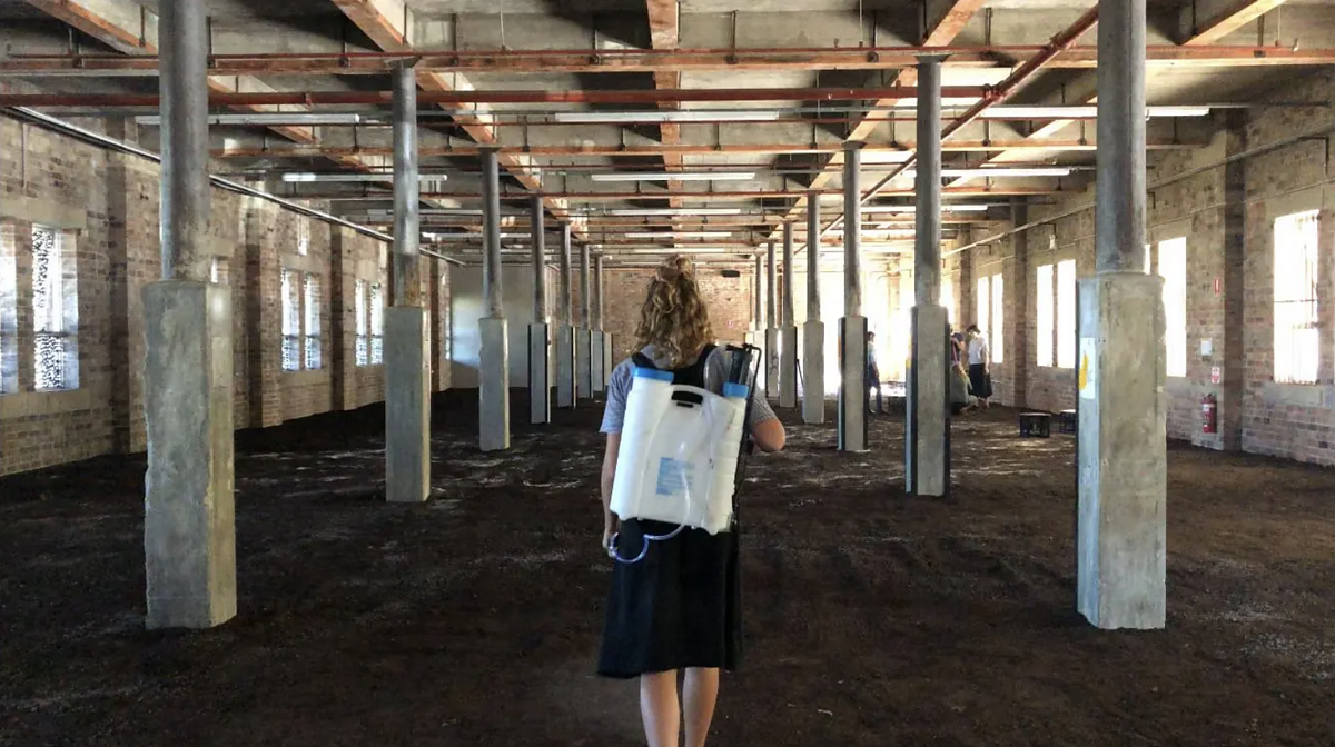 A woman with a backpack stands in an industrial space with soil covering the floor.