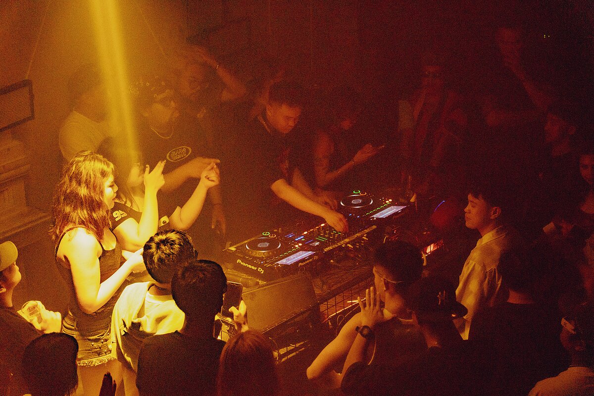 A crowd of people surround a DJ booth on a dimly lit dance floor.