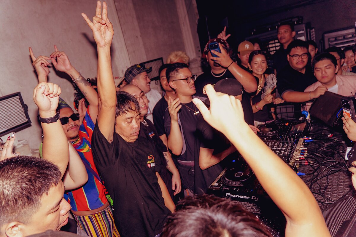 A crowd of people around a DJ booth with their hands in the air.