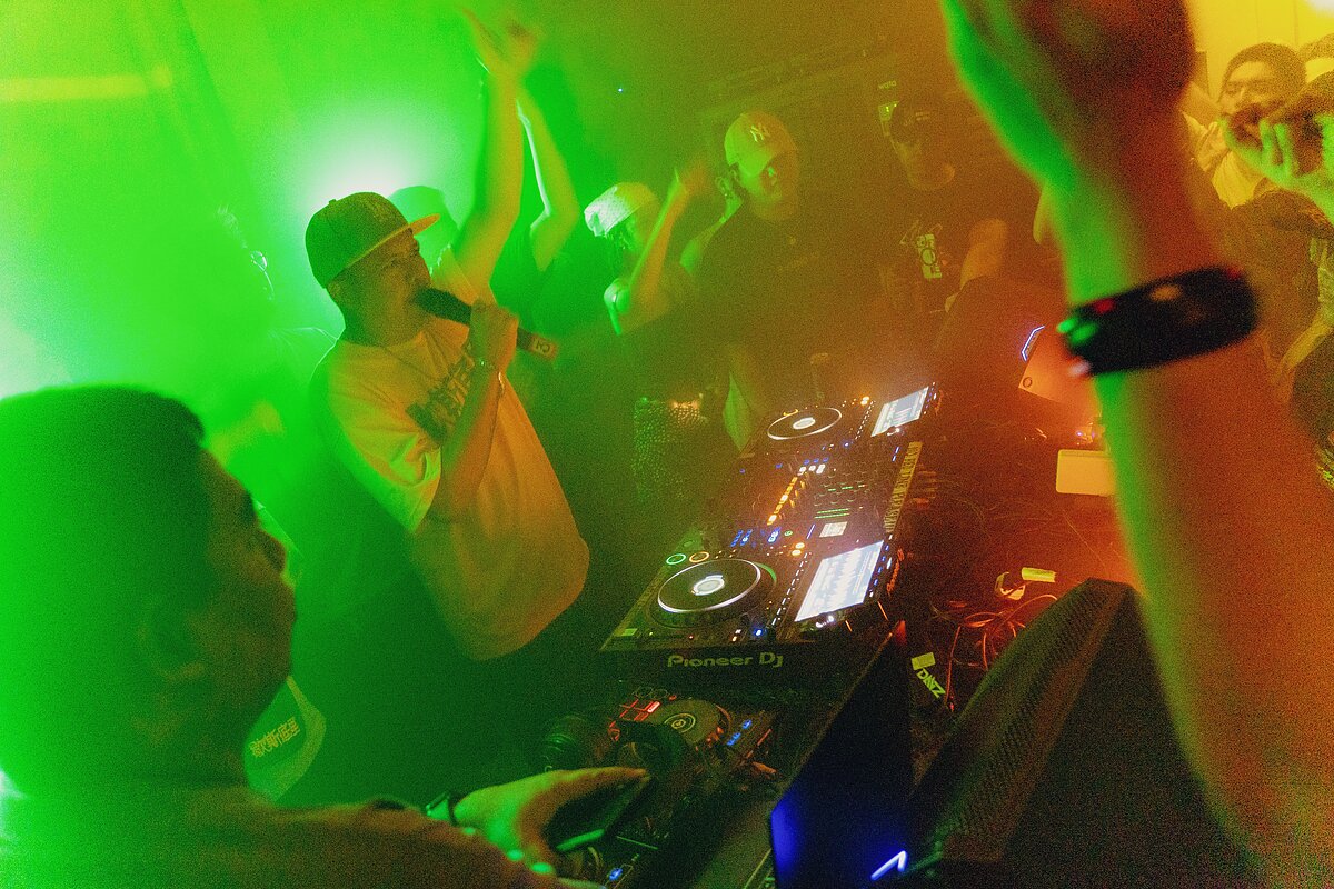 Lit by neon green and orange lights, a man behind a DJ booth holds a microphone while waving one arm in front of a crowd.