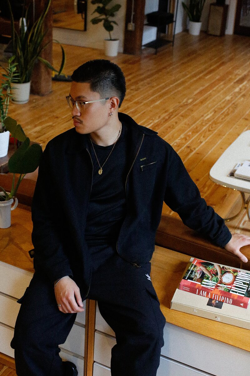 A vertical portrait of a man wearing glasses and all black sitting on wooden furniture in an apartment looks away from the camera.