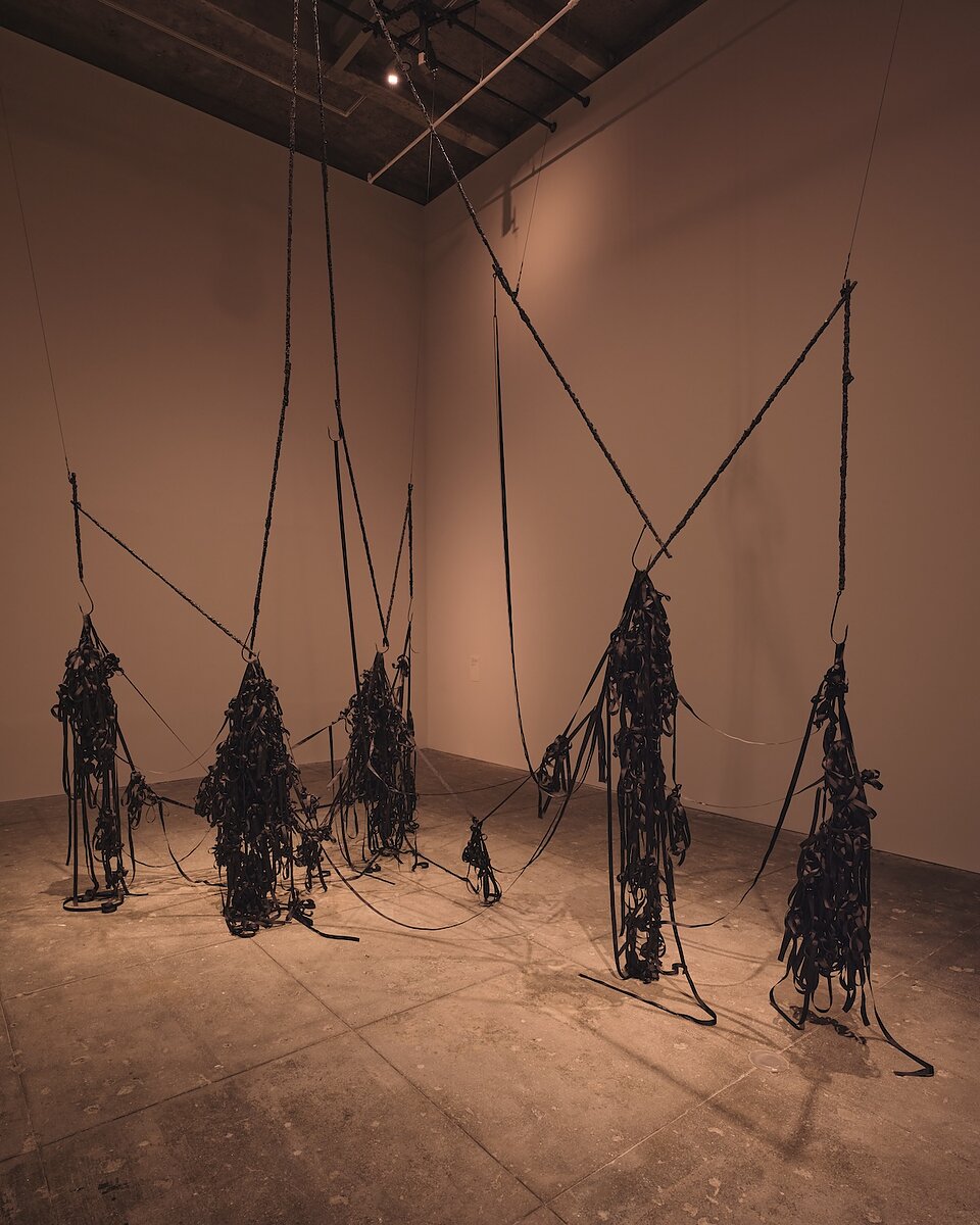A gallery with high ceilings featuring a work of art composed of tangled black fabric hanging from the rafters precariously by sharp hooks.