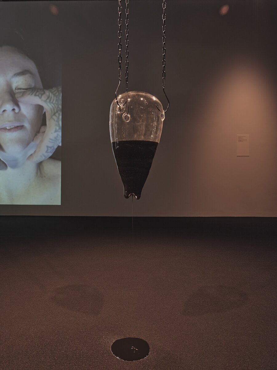A gallery featuring a glass sculpture hanging from the ceiling and dripping a black ooze onto the floor. On the wall behind it is a projected image of a woman's face being massaged by tattooed hands.